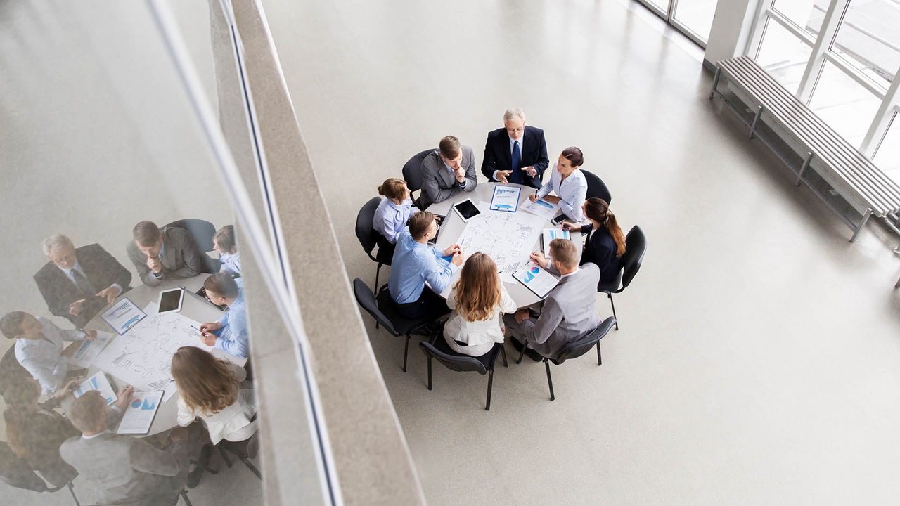A group of professionals discussing around a table in a bright, modern office space. Papers, tablets, and natural light are visible.