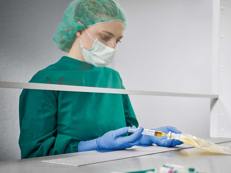 Pielęgniarka w sterylnym kombinezonie z maską ochronną przygotowująca żywienie pozajelitowe w sterylnym środowisku, wstrzykuje płyn do worka żywieniowego. Na pierwszym planie widoczne są różne produkty medyczne.