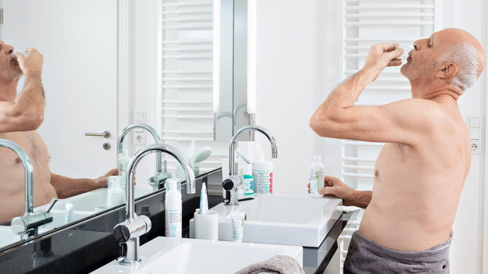 starszy mężczyzna wykonujący płukanie jamy ustnej środkiem przeciwdrobnoustrojowym w domu przed lustrem kąpielowym 