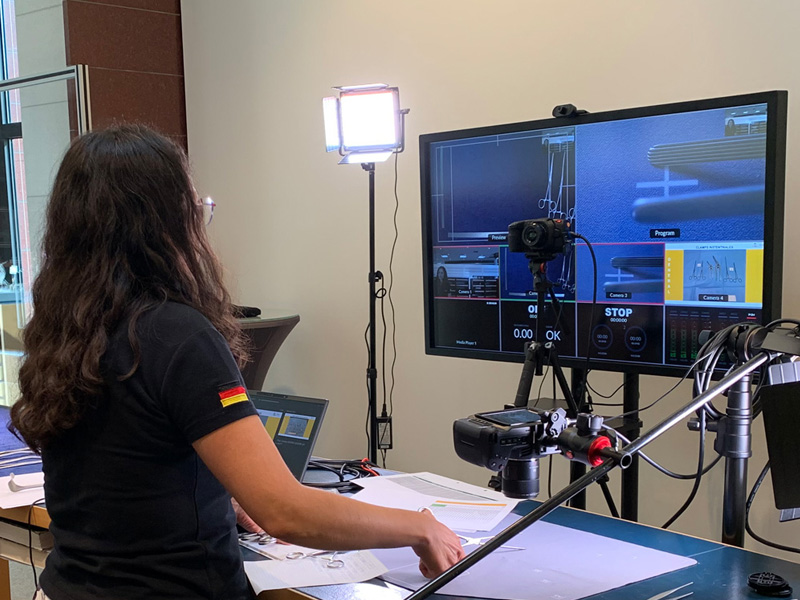 kobieta stojąca przy biurku, na którym znajdują się dwa monitory, statyw z mikrofonem, oświetlenie i kamera